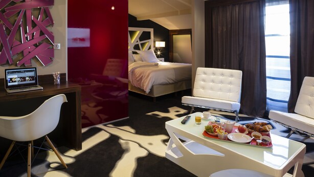 Una habitación de hotel con cama, escritorio con una computadora portátil, mesa de desayuno, sillón blanco y un ventanal con cortinas.
