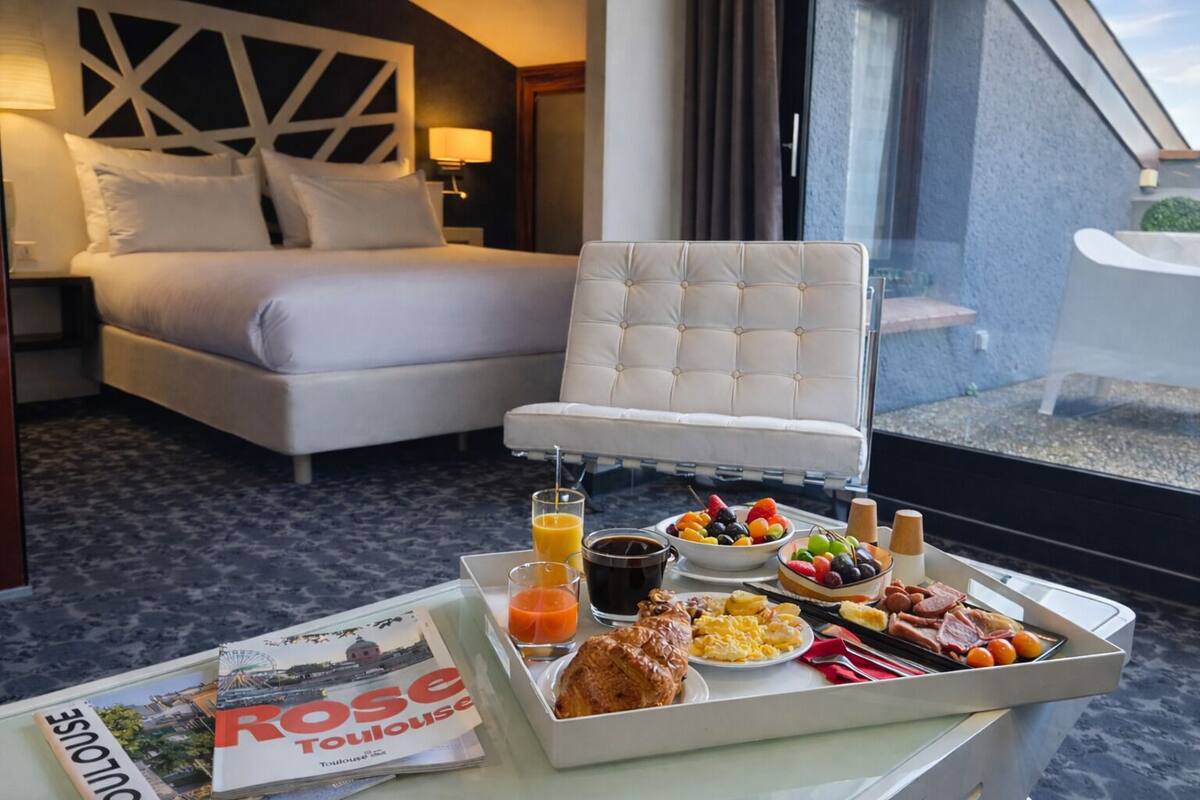 A hotel room with a bed, a white armchair, a breakfast tray with fruits, coffee, and pastries, and a magazine on the table.