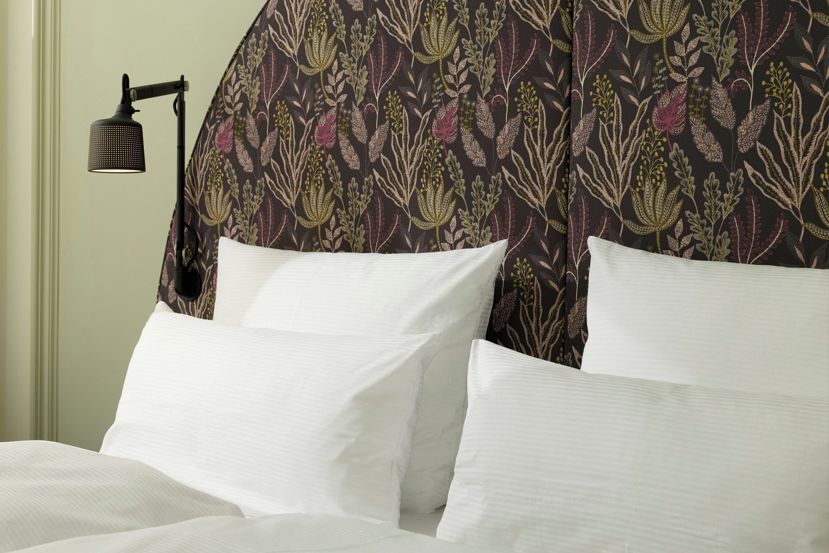 A bed with white linens, a patterned headboard, and a wall-mounted lamp.