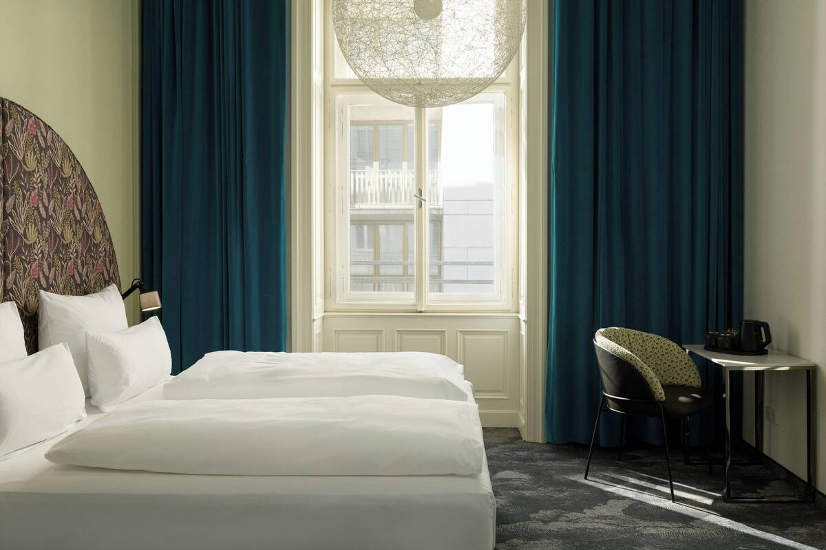 Habitación de hotel con cama, escritorio, silla, ventana con cortinas e iluminación decorativa en el techo.
