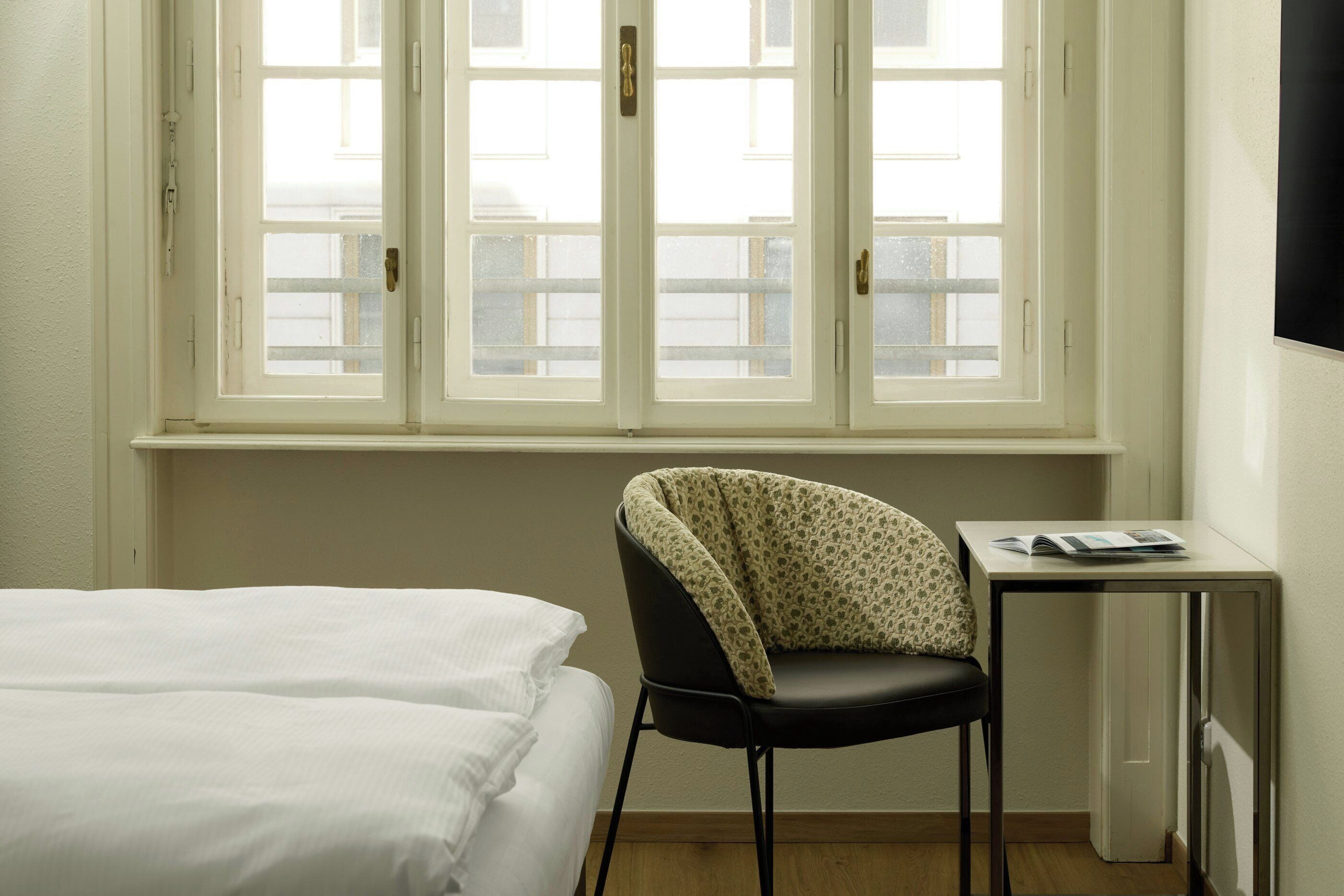 Ein Hotelzimmer mit einem Bett, einem Sessel, einem kleinen Tisch und einem Fenster mit Blick auf Gebäude.
