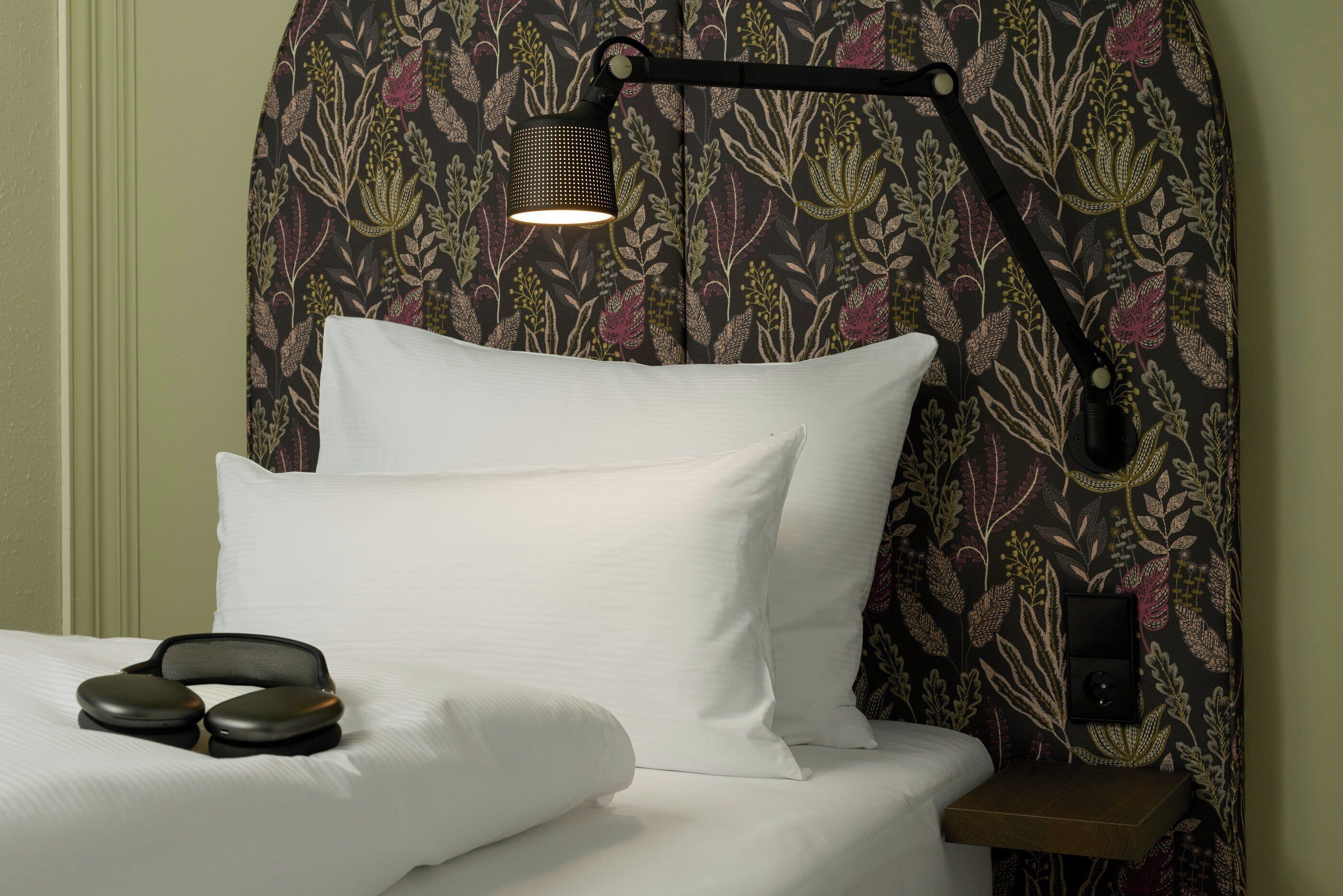 Una cama con ropa de cama blanca, un cabecero con estampado floral, una mesita de noche y una lámpara.