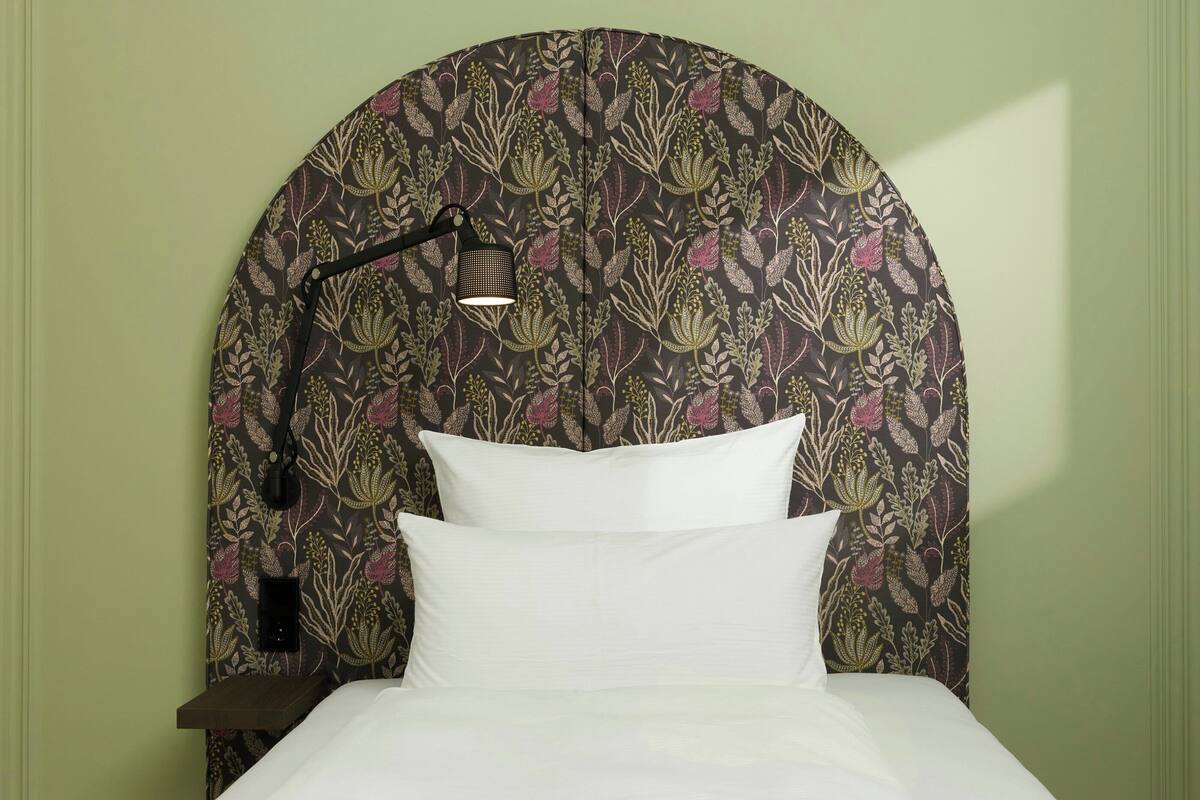 Una cama con ropa blanca, un cabecero con estampado floral y una lámpara de noche.
