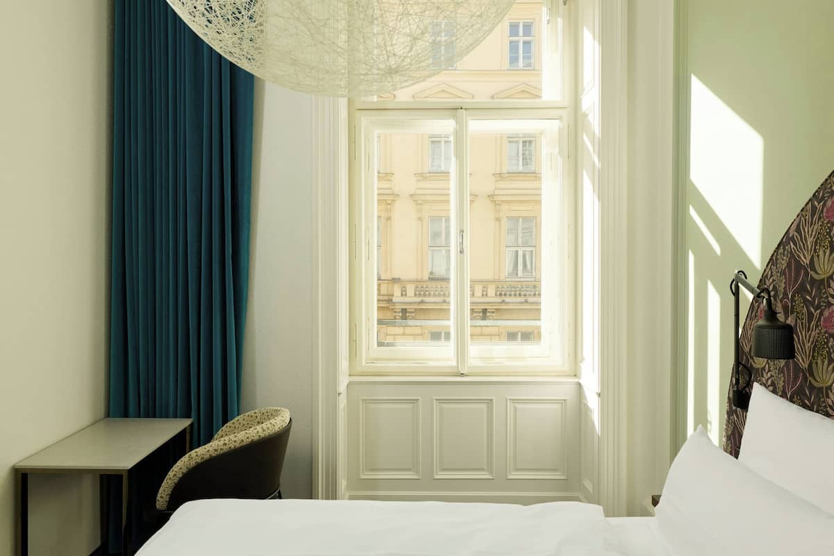 A hotel room with a bed, a desk, a chair, a window with a view of buildings, and a patterned wall lamp.