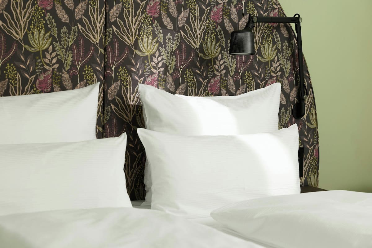 A bed with white linens, a floral-patterned headboard, and a black wall-mounted lamp.