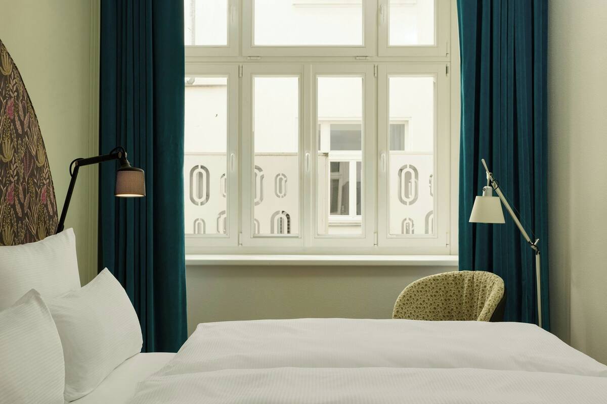 Una habitación de hotel con cama, dos lámparas, una silla y un ventanal con cortinas.