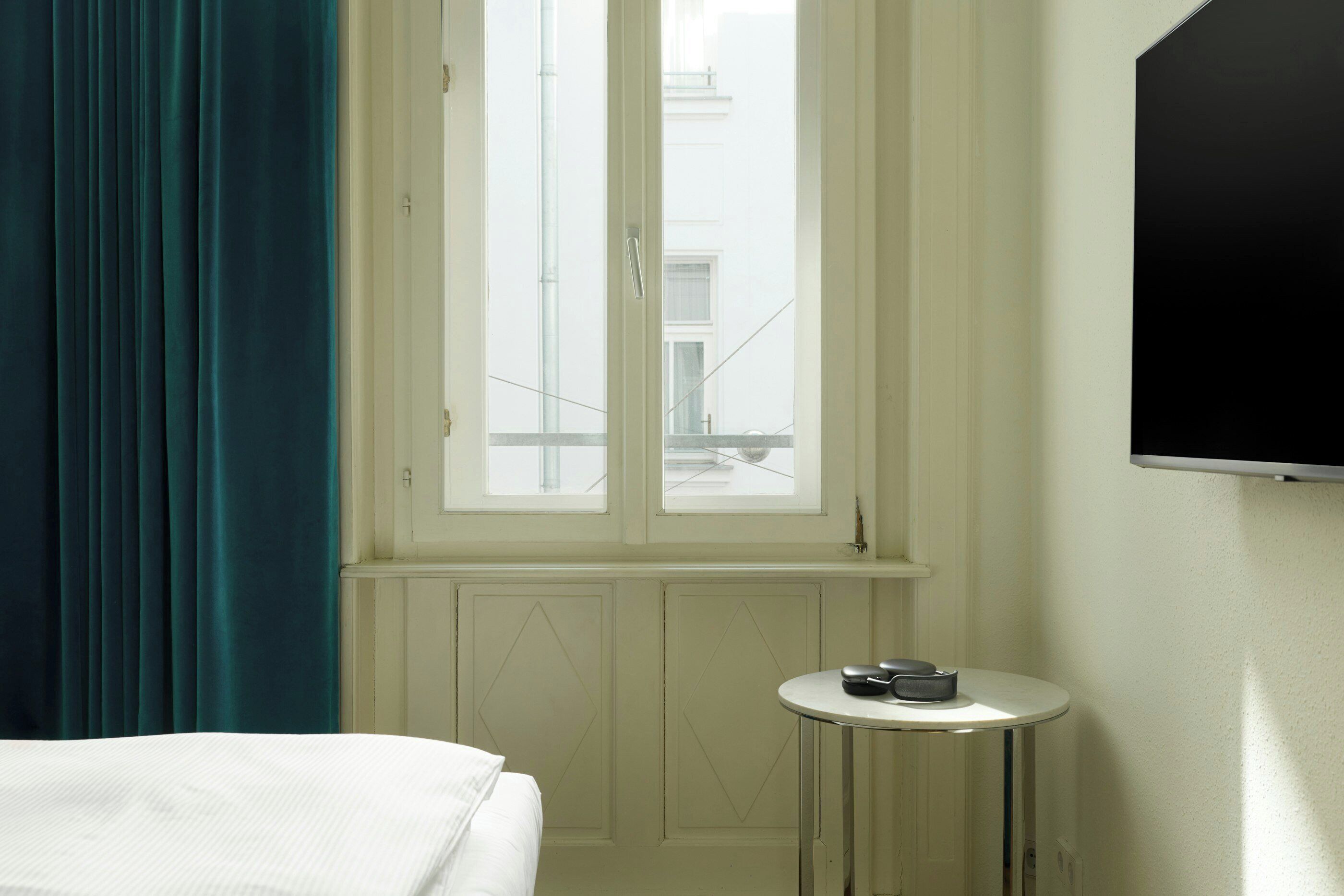 Hotelkamer met een bed, een klein rond tafeltje met een afstandsbediening, een raam met gordijnen en een aan de muur bevestigde televisie.