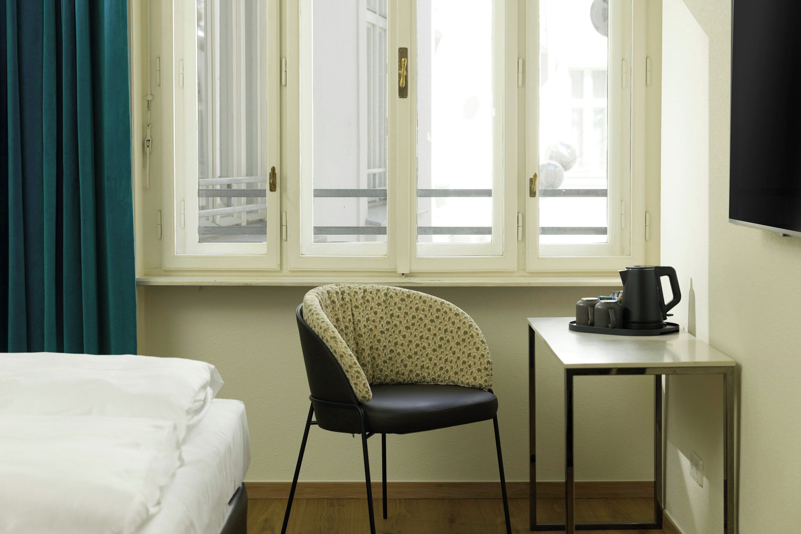 A hotel room with a bed, a chair, a small table with a coffee pot, and a window with a view of a balcony.