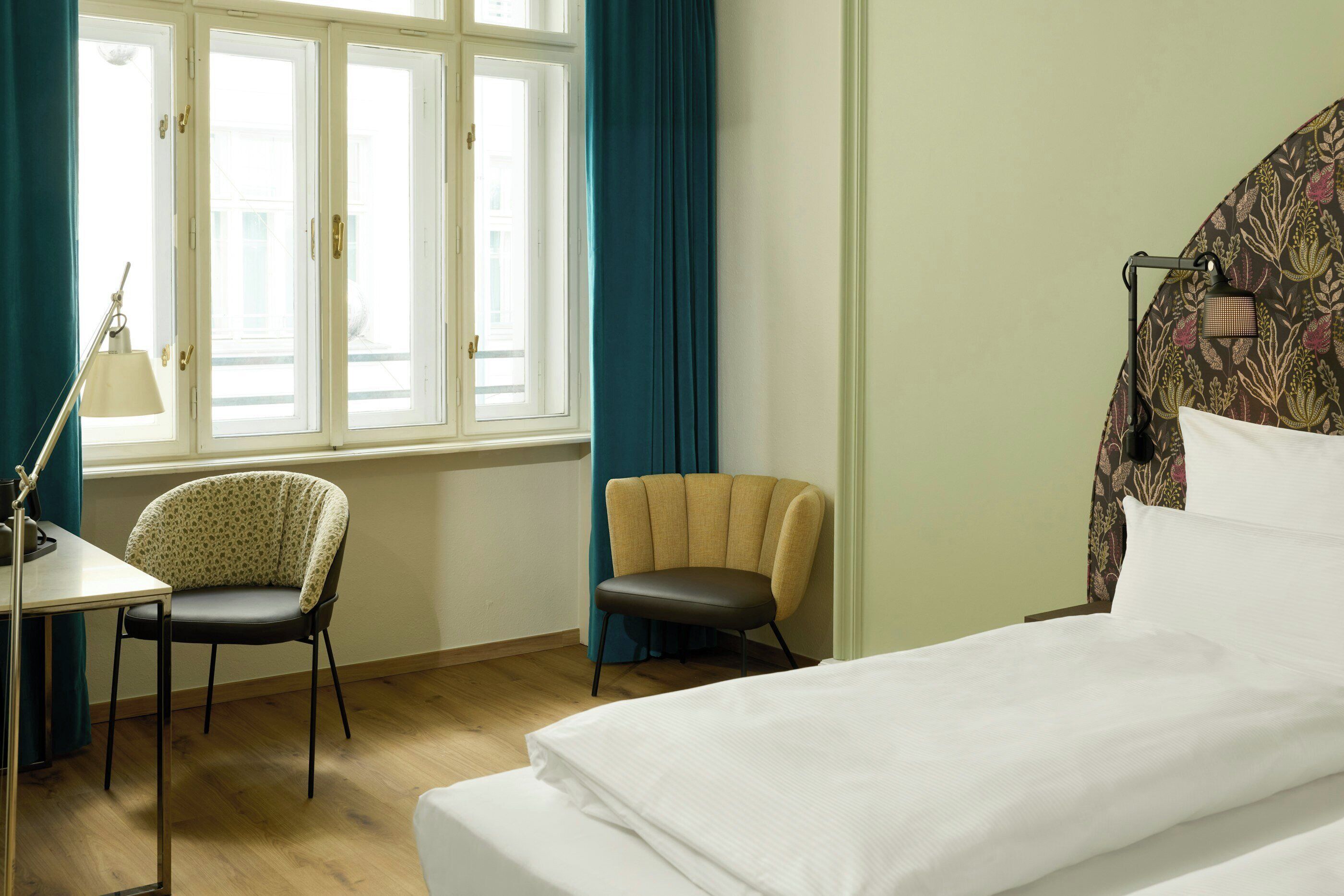 A hotel room with a bed, two chairs, a desk, and a window with curtains.