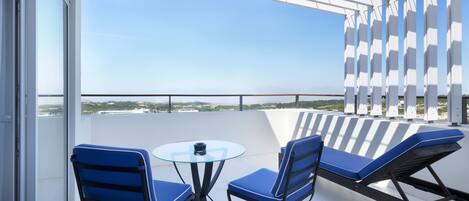 A balcony with a glass table and chairs, a lounge chair, and a view of the cityscape.
