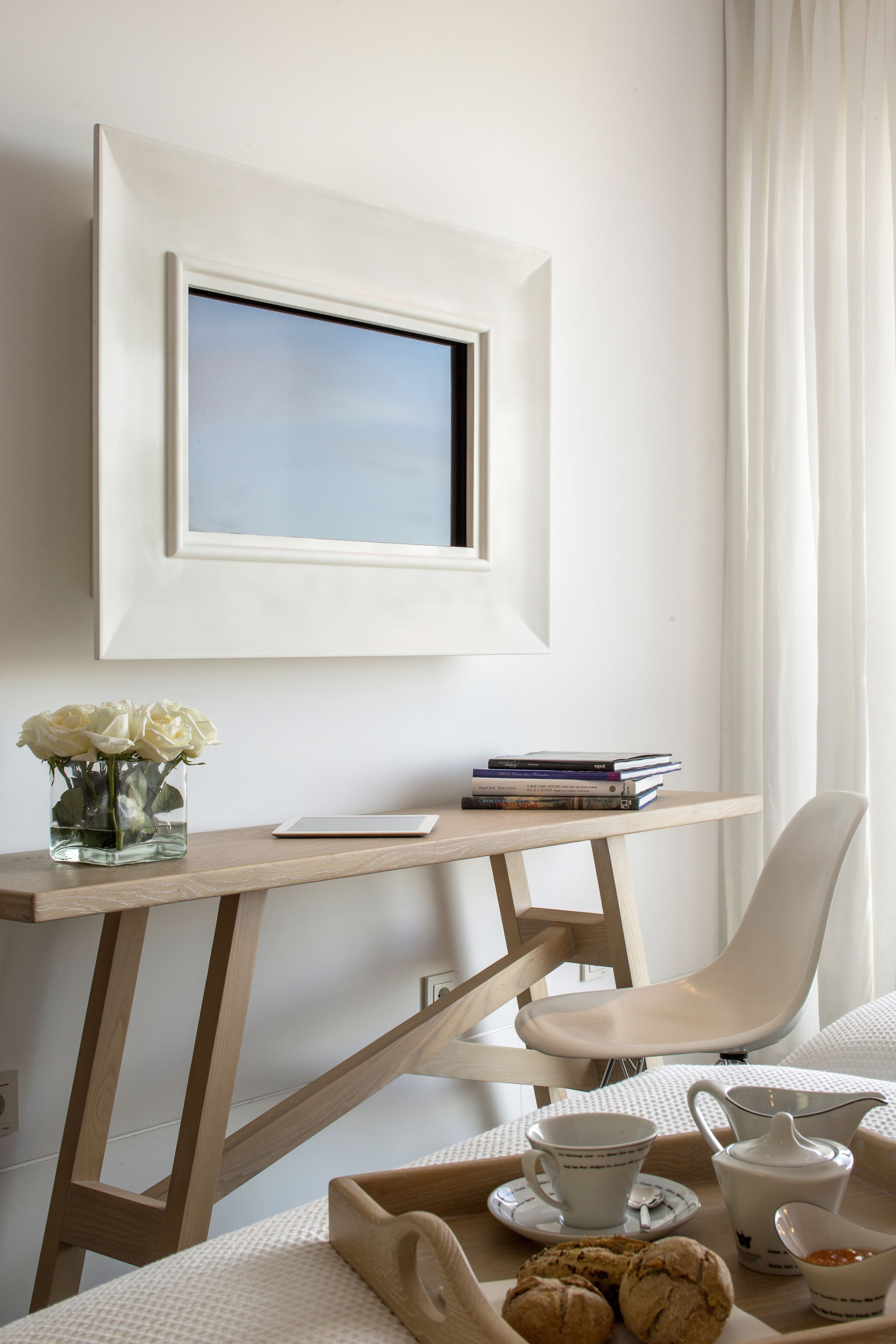 A minimalist room with a desk, a chair, a framed picture, a vase with flowers, and a tray with a cup, teapot, and cookies.