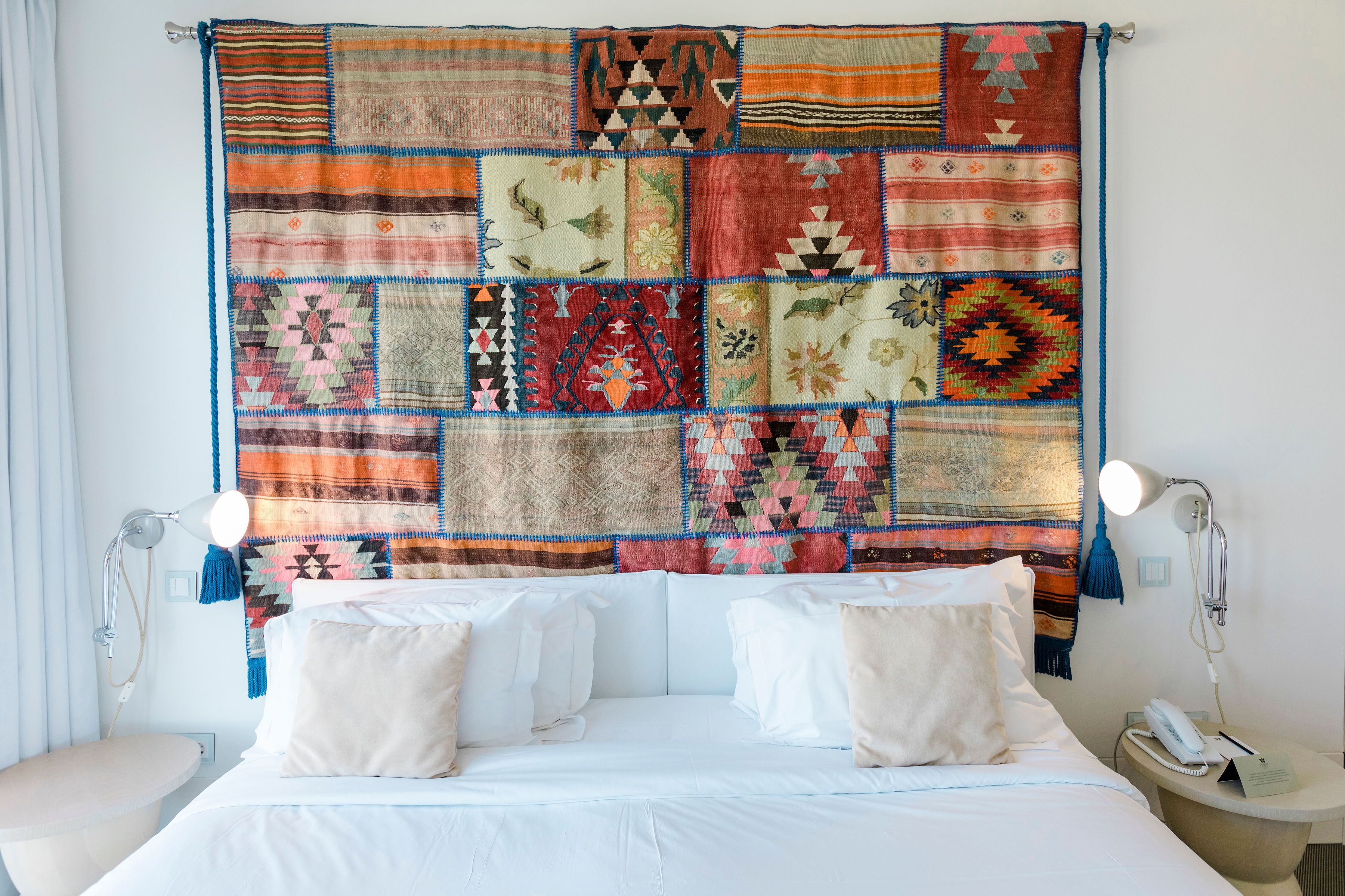 A bed with white linens and pillows, a bedside table with a lamp, and a wall tapestry.