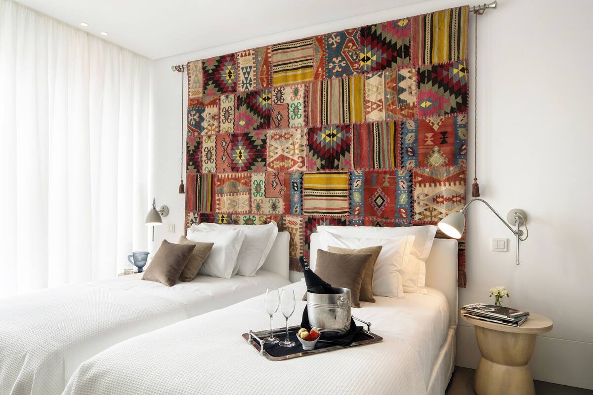 A hotel room with two beds, a large patterned tapestry on the wall, a bedside table with a lamp, and a small round table with a magazine and flowers.