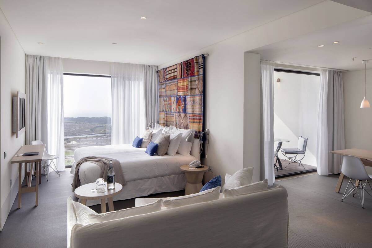 A modern hotel room with a large bed, a small round table, a desk, and a view of the city.