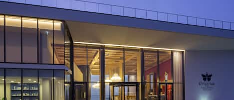 Un hotel moderno con fachada de vidrio, un interior visible con un candelabro y un logotipo en la pared.