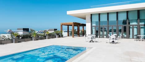 A rooftop pool area with a clear view of the ocean, a covered seating area, and a modern building with large windows.
