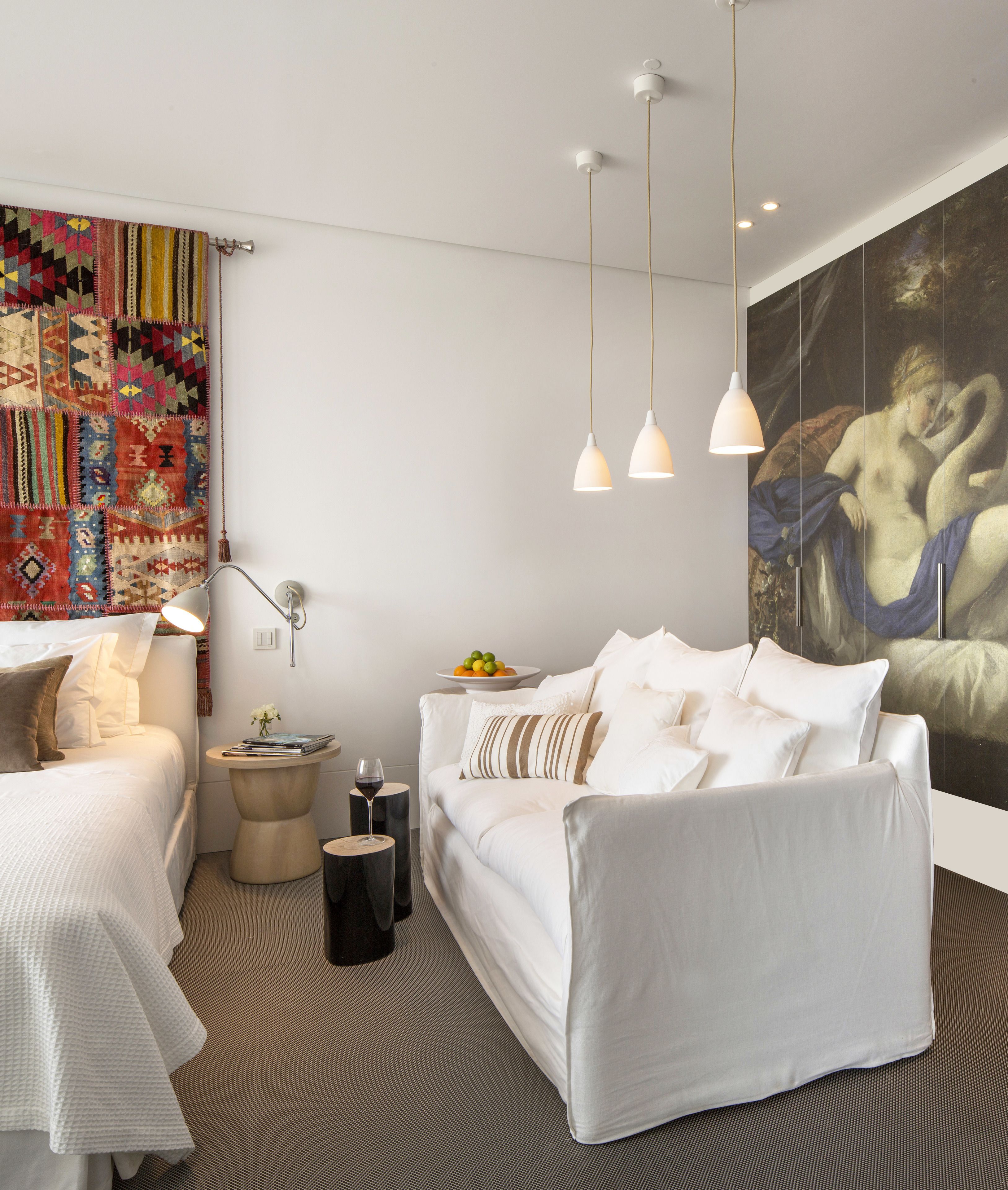 Une chambre moderne avec un lit blanc, un canapé blanc, une petite table ronde avec un verre à vin et une tapisserie colorée au mur.