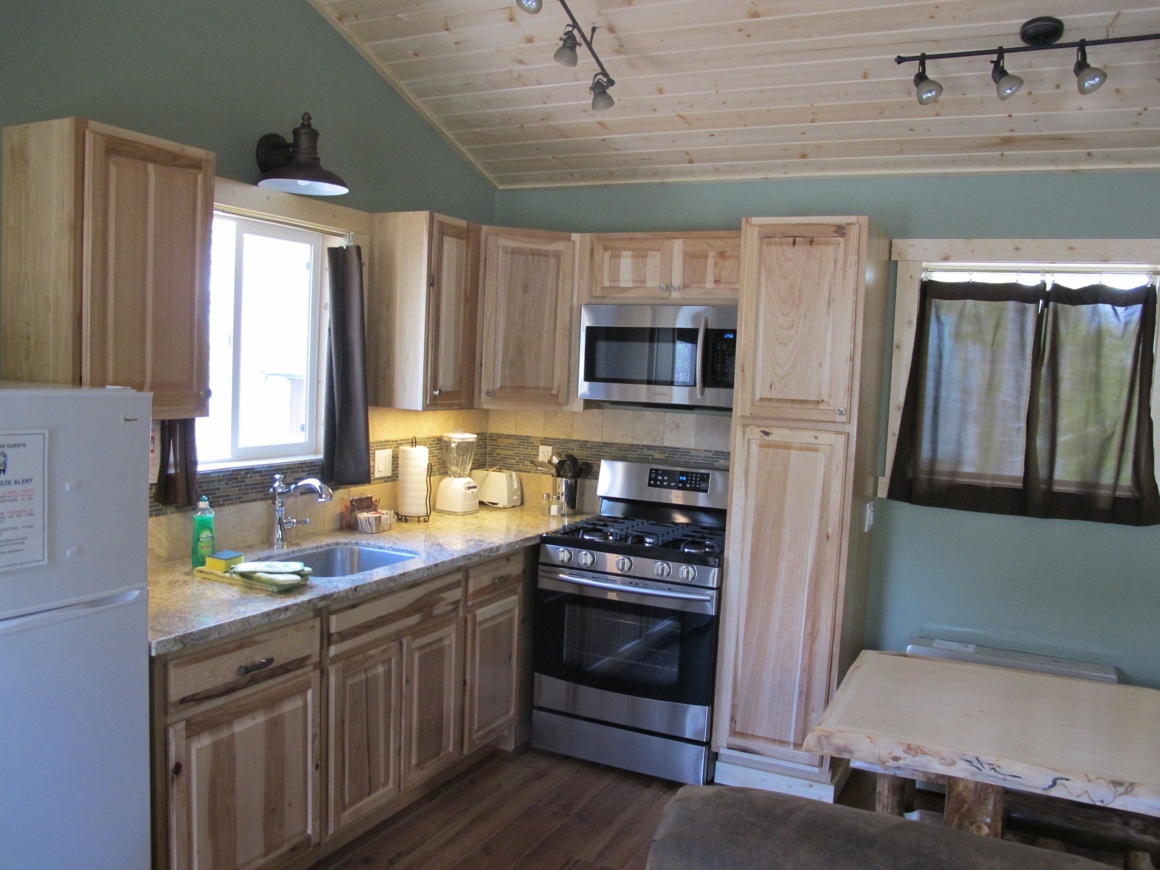 Cocina con armarios de madera, microondas, horno, lavaplatos y ventana con cortinas.