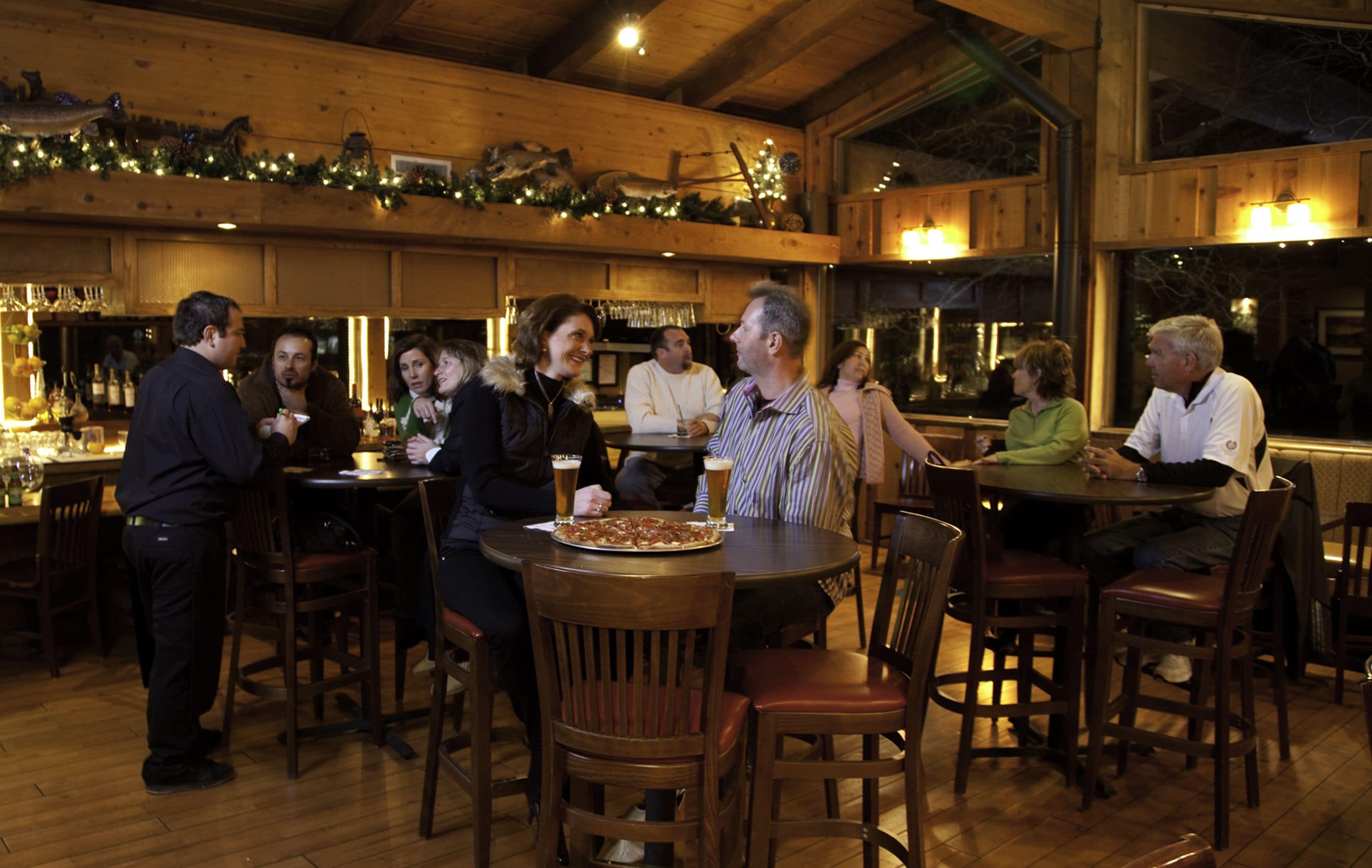 A wooden interior with round tables and chairs, a pizza on one table, and a bar area with bottles and glasses.