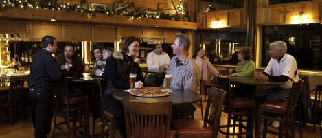 A wooden interior with round tables and chairs, a pizza on one table, and a bar area with bottles and glasses.