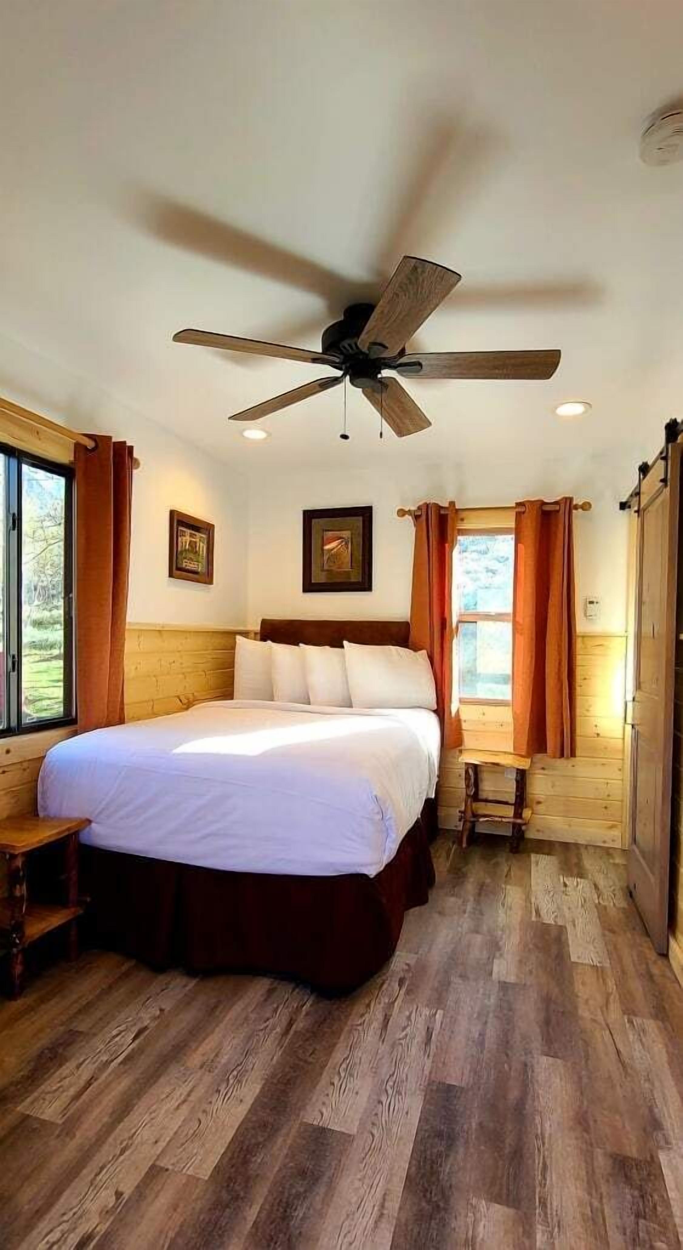 Ein Schlafzimmer mit Bett, Holzboden, Deckenventilator und Fenster mit Vorhängen.