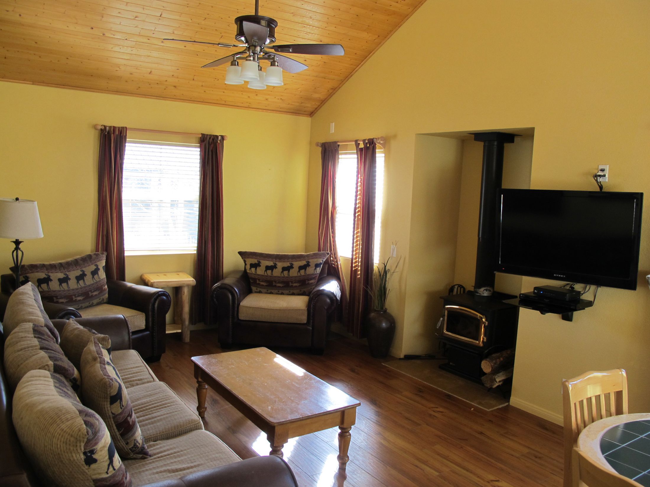 A living room with a wooden ceiling, a ceiling fan, a flat-screen TV mounted on the wall, a wooden coffee table, a sofa with patterned cushions, and a window with curtains.