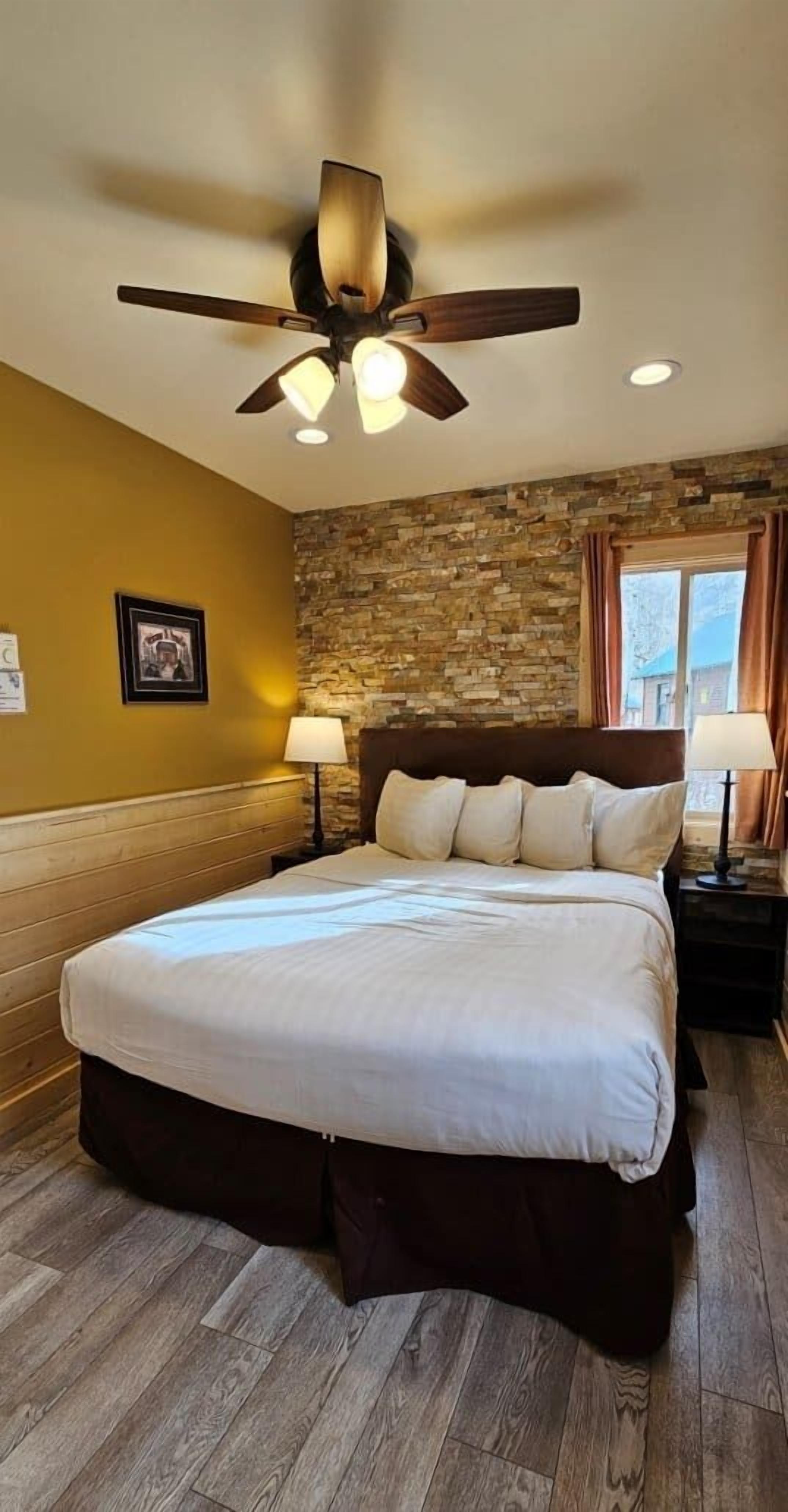 A bedroom with a large bed, a ceiling fan, a stone wall, and a window with curtains.