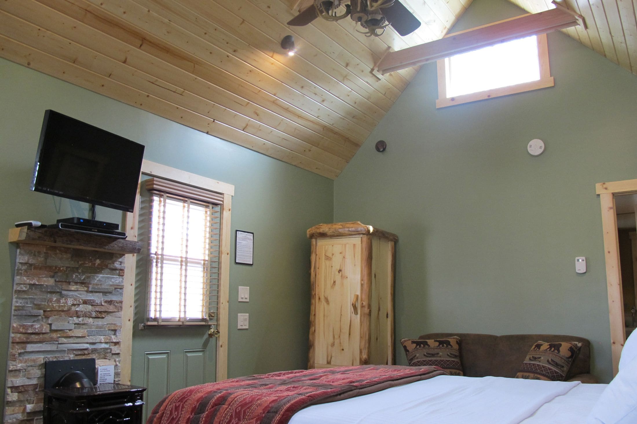 Een slaapkamer met een houten plafond, een aan de muur bevestigde televisie, een stenen openhaard, een houten garderobe en een bed met gekleurde kussens.