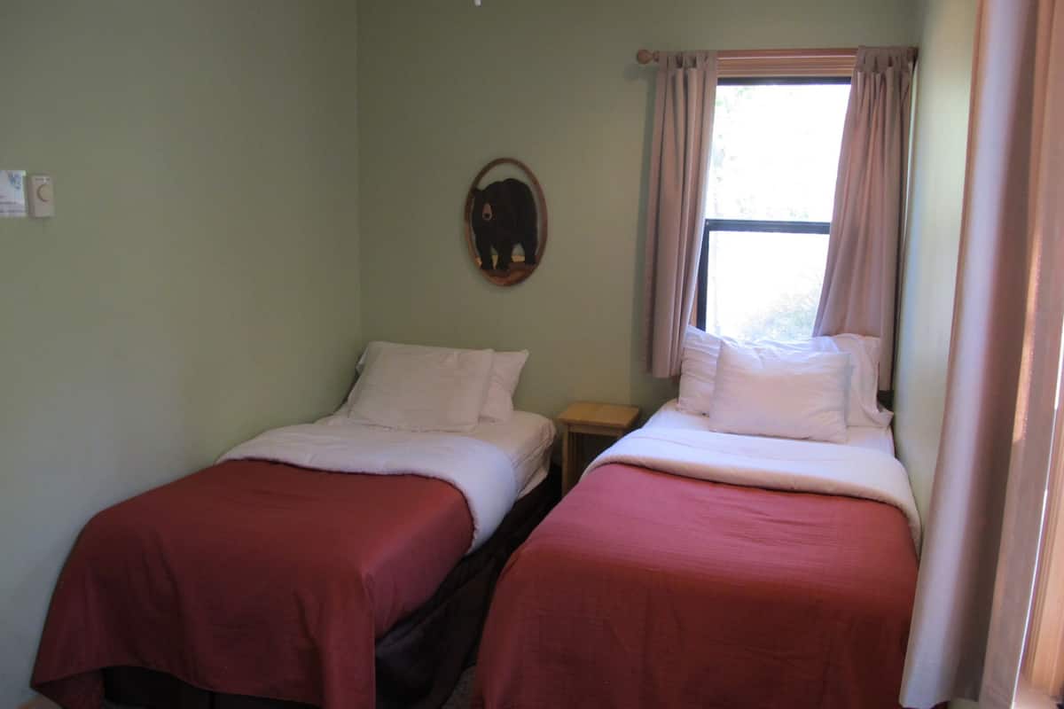 A room with two beds, each with a red bedspread and white pillows, a small wooden nightstand, a wall-mounted mirror, and a window with curtains.