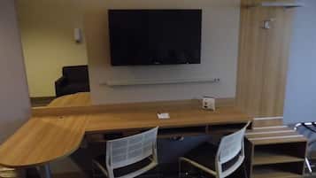 A hotel room with a wooden desk, two white chairs, a flat-screen TV mounted on the wall, and a small shelf below the TV.