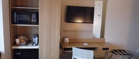 A compact kitchenette with a microwave, coffee maker, and a small refrigerator. A flat-screen TV is mounted above a wooden desk, and a chair is placed beside it.