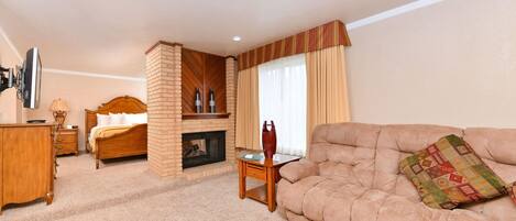 A hotel room with a bed, a sofa, a wooden dresser, a small table, a fireplace, and a television.