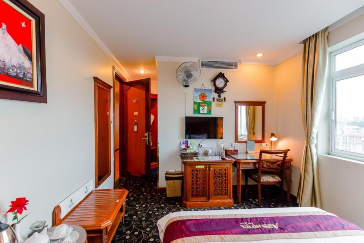 Habitación de hotel con cama, escritorio, silla, televisión, reloj, ventilador y ventana.