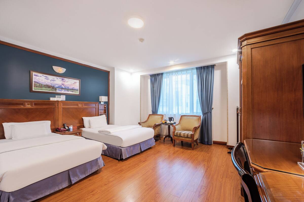 A hotel room with two beds, a wooden wardrobe, a desk with a chair, a small table with a lamp, and a window with curtains.