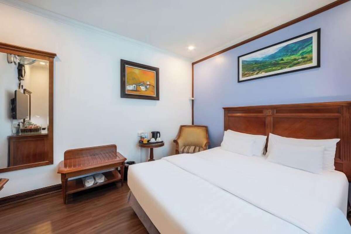 A hotel room with a large bed, a wooden bench, a small table with a kettle, and two framed pictures on the walls.