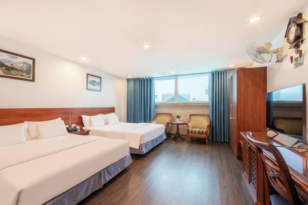 A hotel room with two beds, a desk with a TV, a chair, a small table with a plant, and a window with curtains.