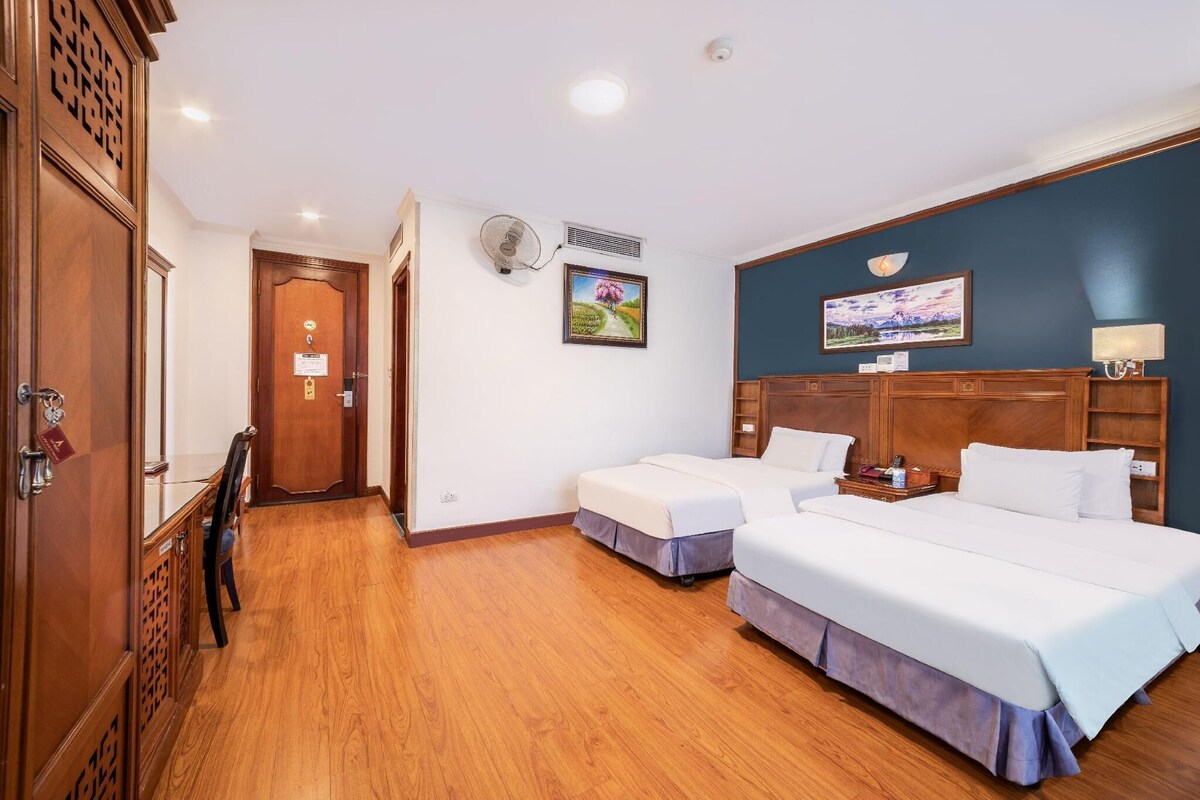 A hotel room with two beds, a wooden wardrobe, a desk, a chair, and a wall-mounted fan.