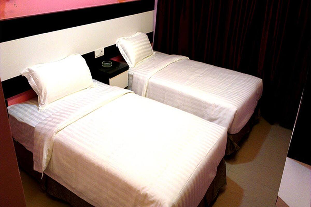 A hotel room with two single beds, each with white linens and pillows, a nightstand with a lamp, and a dark curtain in the background.