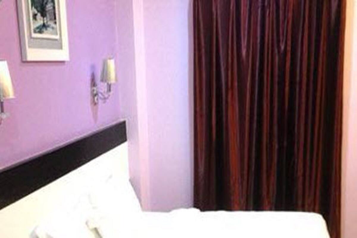 A neatly made bed with white linens in a room with purple walls and a dark curtain.