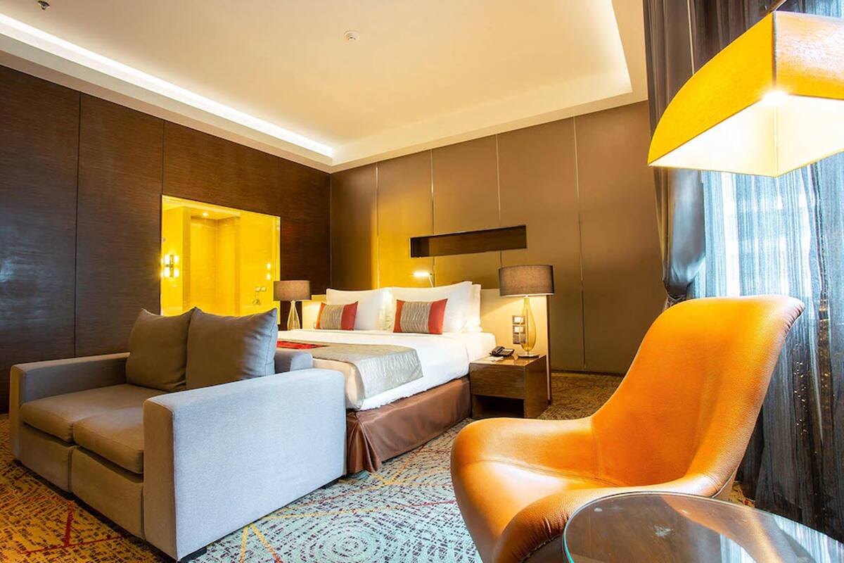 Une chambre d’hôtel avec un lit, un canapé, une chaise, une table de chevet et une lampe.