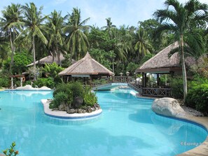 A resort pool with thatched-roof structures, surrounded by palm trees and tropical vegetation. - Coco Beach Island Resort (Puerto Galera)
