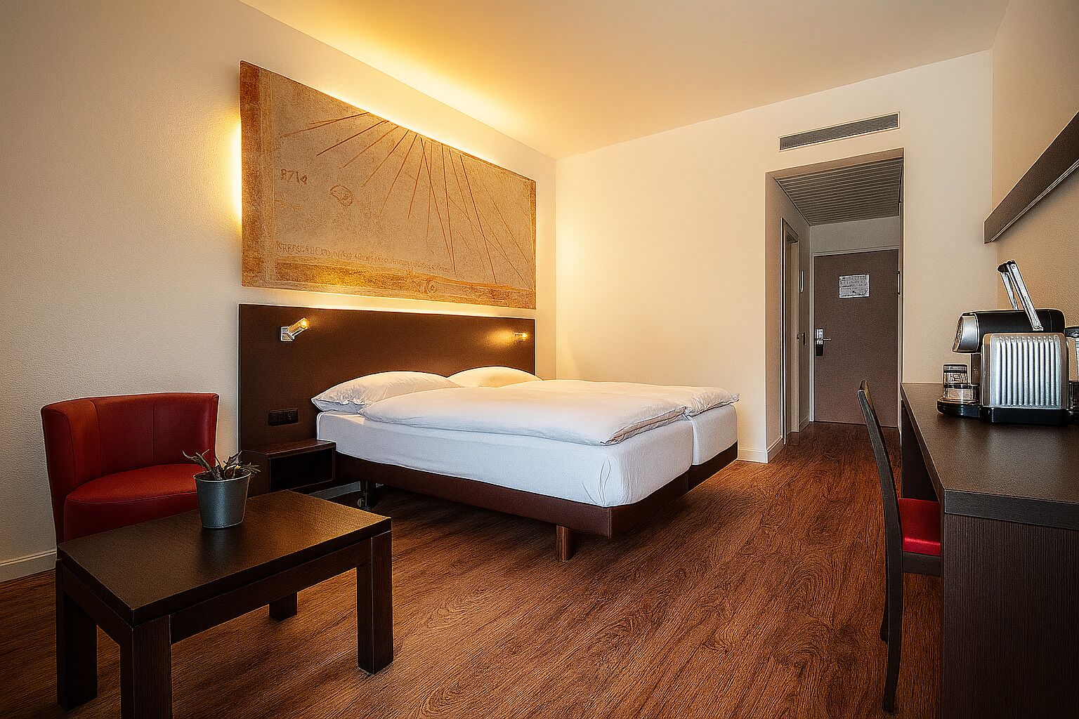 Una habitación de hotel con una cama, una silla roja, una mesita pequeña y un escritorio con un radio.