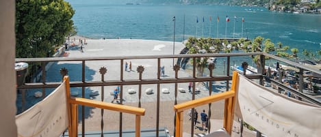 A balcony with two white cushioned chairs, a railing, and a view of a beach and lake with mountains in the background.
