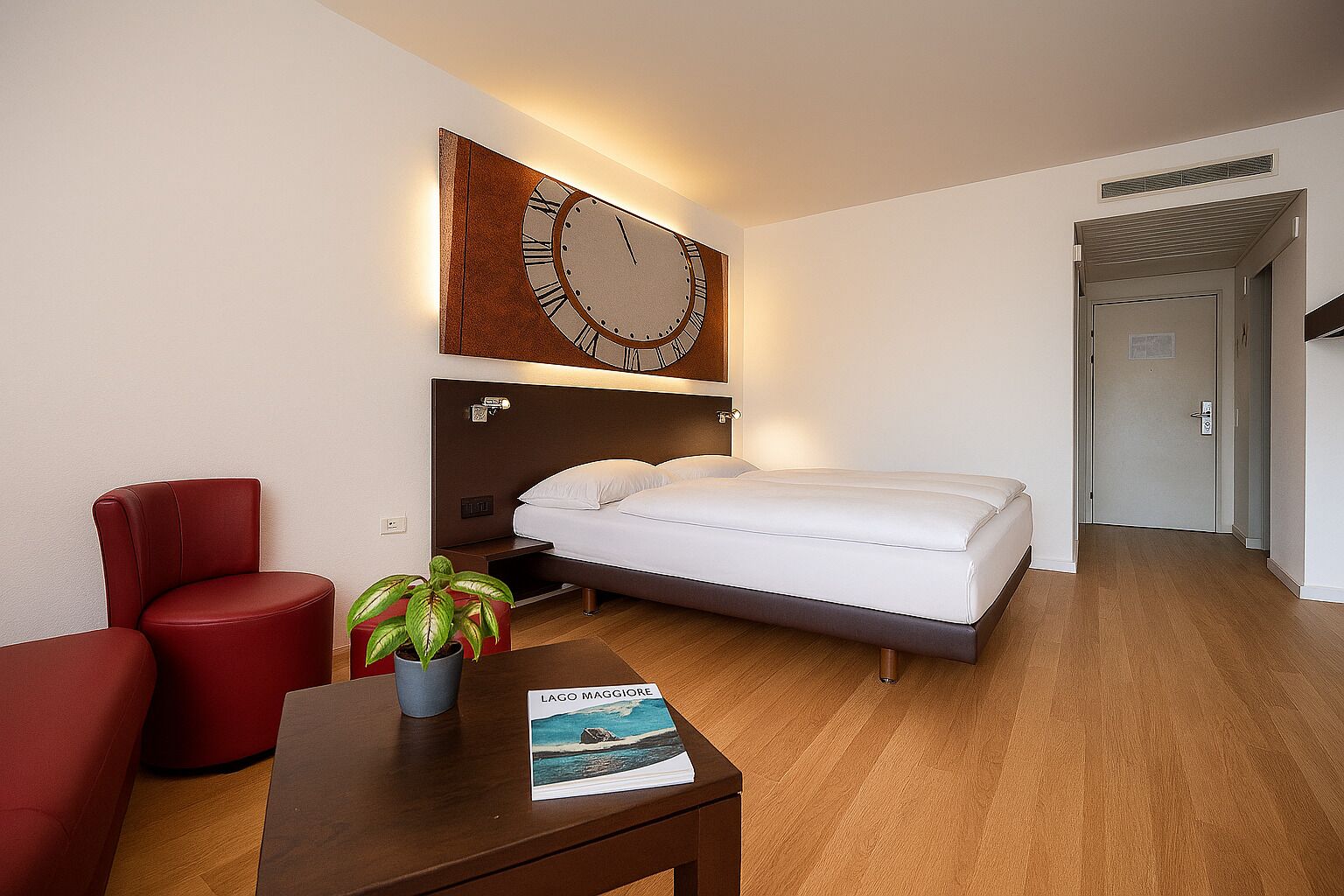 Una habitación de hotel con una cama, un sillón rojo, una mesita con un libro y un reloj de pared.
