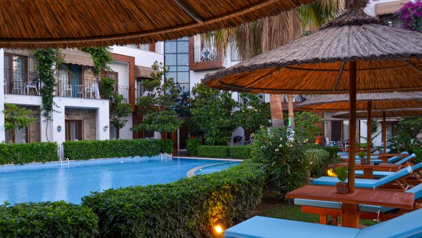 A pool area with lounge chairs and a thatched umbrella, surrounded by greenery and a building with balconies. - Liona Hotel (Bodrum)