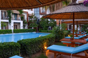 A pool area with lounge chairs and a thatched umbrella, surrounded by greenery and a building with balconies. - Liona Hotel (Bodrum)