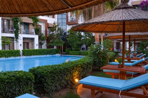 Un área de piscina con sillas de descanso y una sombrilla de paja, rodeada de follaje y un edificio con balcones.