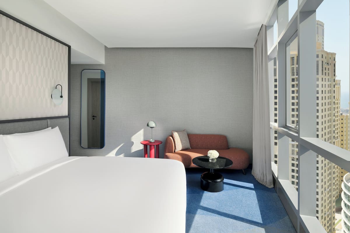 A hotel room with a bed, a sofa, a small round table, a mirror, and a view of the cityscape.