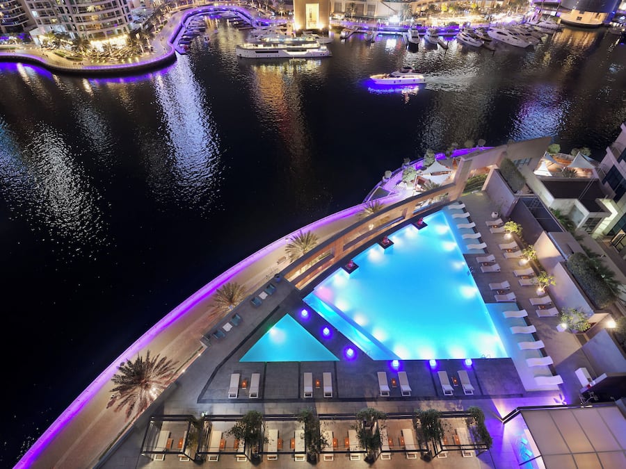 Vista noturna de um hotel à beira-mar, com piscina, áreas de estar e barcos atracados nas proximidades.