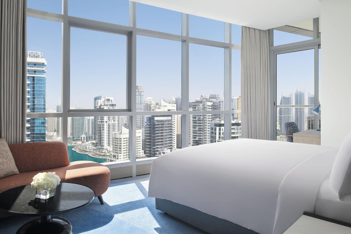 A modern hotel room with a large bed, a sofa, and a coffee table. The room features large windows with a city view.